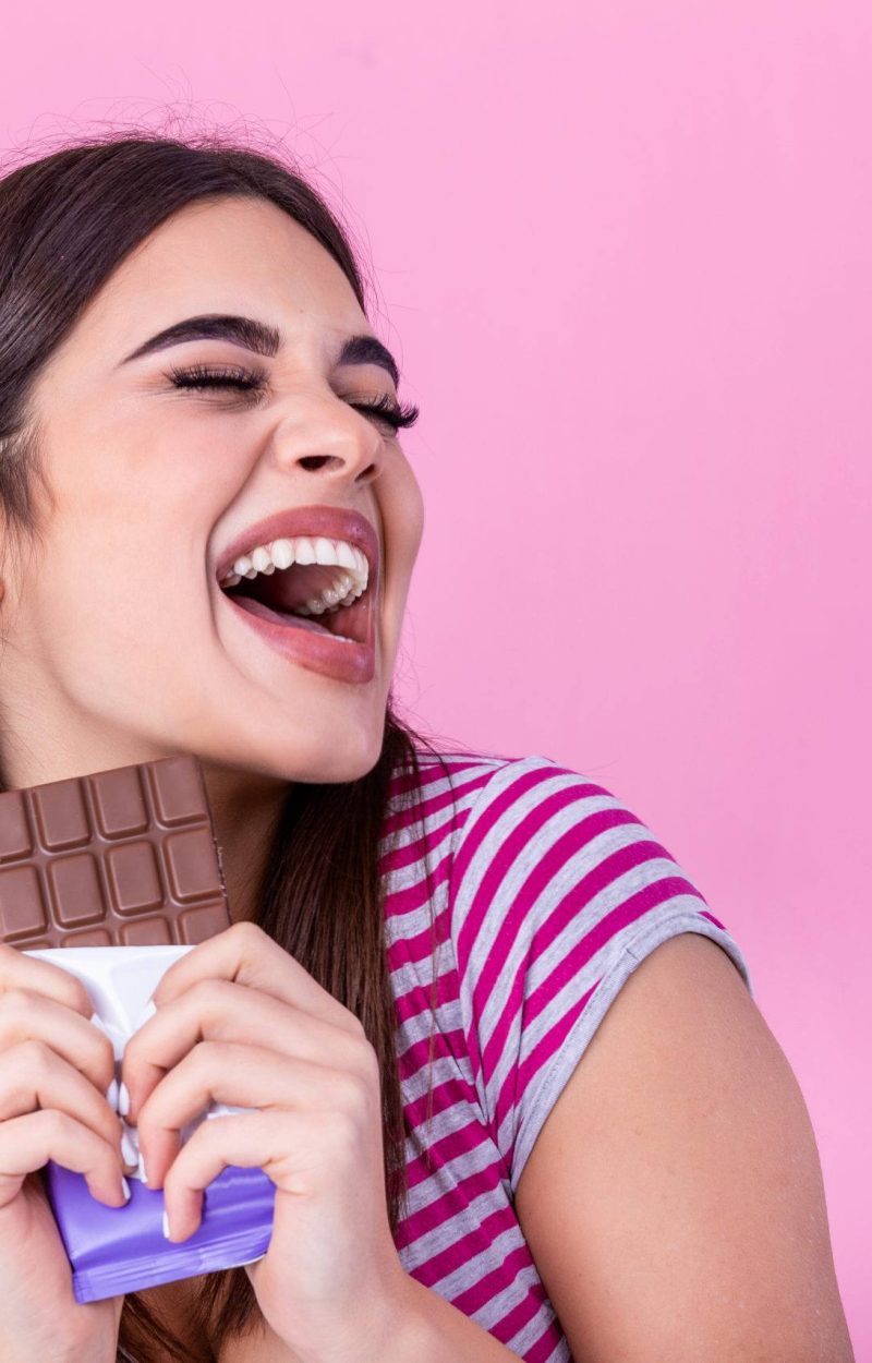 Happy young beautiful lady eating chocolate and smiling. Girl tasting sweet chocolate. Young woman with natural make up having fun and eating chocolate isolated on pink background